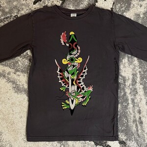 May include: Dark gray long-sleeve shirt featuring a colorful graphic of a dagger piercing a frog with butterfly wings. The design includes red, green, yellow, and white accents. The shirt is laid flat.