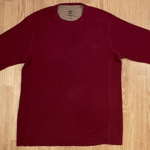 May include: A long-sleeve crew neck shirt in a deep maroon color. The garment features a textured waffle-knit fabric, a solid design, and a contrasting tan interior lining at the collar with a visible Timberland brand logo and text.