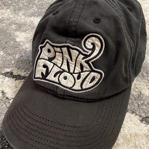 May include: A charcoal gray baseball cap featuring an embroidered patch on the front. The patch is white with a black outline and displays the text Pink Floyd in a stylized font. The cap has a curved brim and stitched eyelets on the crown.