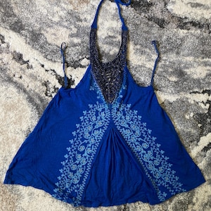 Puede incluir: Un top azul con cuello halter con un escote de cuentas y un diseño floral. El top tiene un escote en V profundo y tirantes finos. La tela es de color azul liso con un estampado floral azul claro.