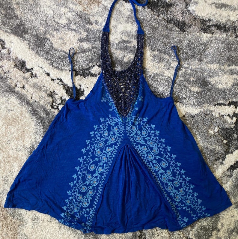 K&ouml;nnte beinhalten: Ein blaues Neckholder-Top mit Perlenbesatz am Ausschnitt und floralem Design. Das Top hat einen tiefen V-Ausschnitt und d&uuml;nne Tr&auml;ger. Der Stoff ist einfarbig blau mit einem helleren blauen Blumenmuster.