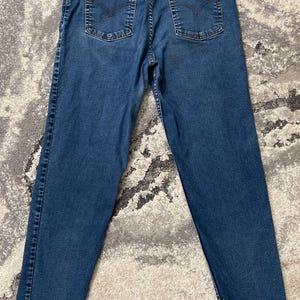 May include: Medium-wash blue denim jeans featuring a high-waisted fit, five-pocket styling, and a tan leather patch on the back waistband. The back pockets include a signature arcuate stitch design and a small red tab with Levi Strauss & Co text.