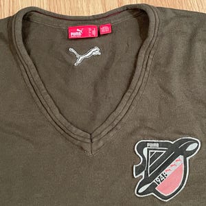 Può includere: Un maglione con scollo a V color verde oliva con un logo Puma bianco ricamato sotto lo scollo. Una grande toppa a forma di scudo è cucita sul lato inferiore destro, riportante la scritta Puma, l'anno 1948 e un grafico rosso con icone puma.