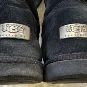 May include: Close-up of two black UGG boots. Each boot features a silver rectangular emblem with the brand name "UGG Australia". The boots have a suede-like texture and a dark sole. The image highlights the brand's logo and the boots' design.