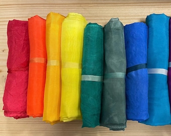 May include: A collection of ten rolled fabric scarves arranged in a spectrum of colors including red, orange, yellow, green, teal, blue, and purple. Each individual scarf is secured with a thin, matching ribbon band wrapped around the center.