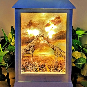 Könnte beinhalten: Eine weiße Laterne mit einer durchsichtigen Glasscheibe, die ein Foto von zwei Giraffen zeigt, die sich in einer Savannenlandschaft küssen. Die Laterne hat ein Licht im Inneren, das das Bild beleuchtet.