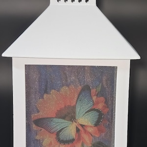 Pode incluir: Lanterna branca com um painel de vidro transparente que apresenta uma borboleta colorida em uma flor. A lanterna tem uma moldura de metal branco e uma alça na parte superior.