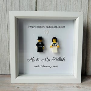 May include: A white framed wedding gift with Lego figures of a bride and groom. The frame includes the text "Congratulations on tying the knot!" and the names "Mr & Mrs Sellick" with the date "20th February 2025".
