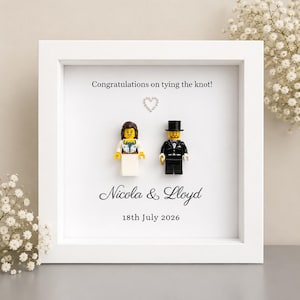 May include: A white square shadow box frame containing two plastic toy figures dressed in wedding attire. The text reads: Congratulations on tying the knot! Nicola & Lloyd 18th July 2026. A small crystal heart is positioned between the figures.