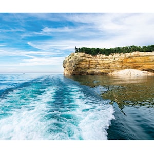 May include: A boat leaves a white foamy wake in the turquoise water of Lake Superior. The boat is traveling towards a large cliff with layered rock formations and green trees on top.