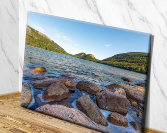 May include: A panoramic view of a lake with mountains in the background. The lake is calm and the water is a light blue color. There are large rocks in the foreground, some of which are partially submerged in the water.