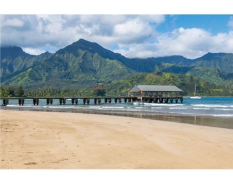 May include: A wooden pier with a roof extends out into the ocean, with a sandy beach in the foreground and a mountain range in the background.