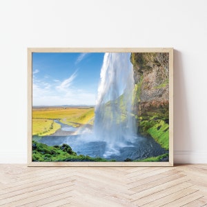 Op de afbeelding: Een ingelijste foto van een waterval die over een rotsachtige klif stort. Het water is melkachtig wit en de klif is bedekt met groen mos. De lucht is blauw met witte wolken.