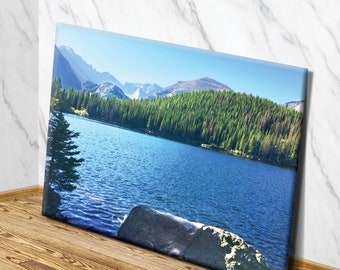 May include: A scenic landscape photograph featuring a lake surrounded by a dense forest of evergreen trees. The lake is a deep blue color and the water is calm and still. The mountains in the background are covered in snow and the sky is a clear blue.