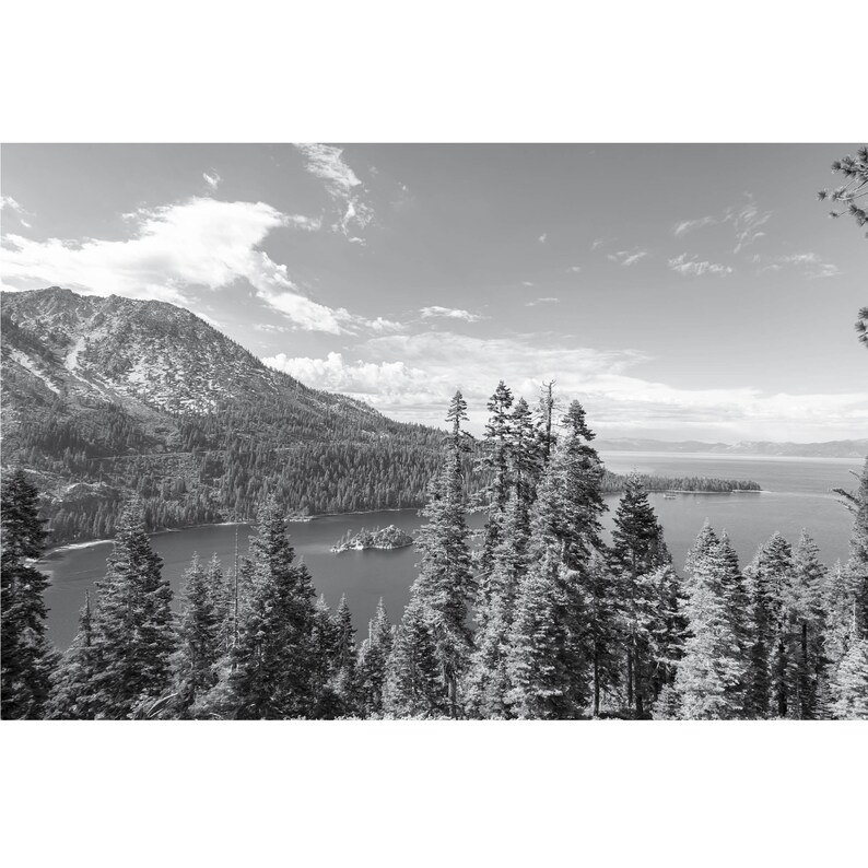 May include: A black and white photograph of a mountain lake with a small island in the middle. The lake is surrounded by trees and mountains. The sky is cloudy.