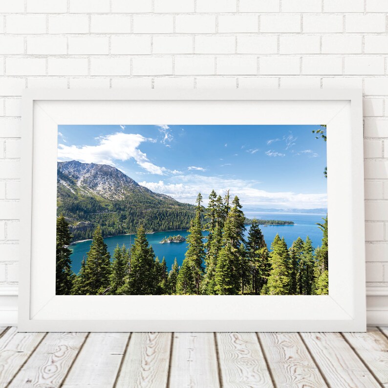 May include: A framed photograph of a scenic view of a lake surrounded by mountains and trees. The lake is a bright blue color and the mountains are covered in trees and snow. The sky is a clear blue with white clouds.