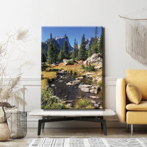 May include: A scenic landscape photograph featuring a mountain range with a stream flowing through a valley. The stream is surrounded by lush green trees and rocks. The mountains are in the background and are covered in trees. The sky is blue and there are clouds in the sky.