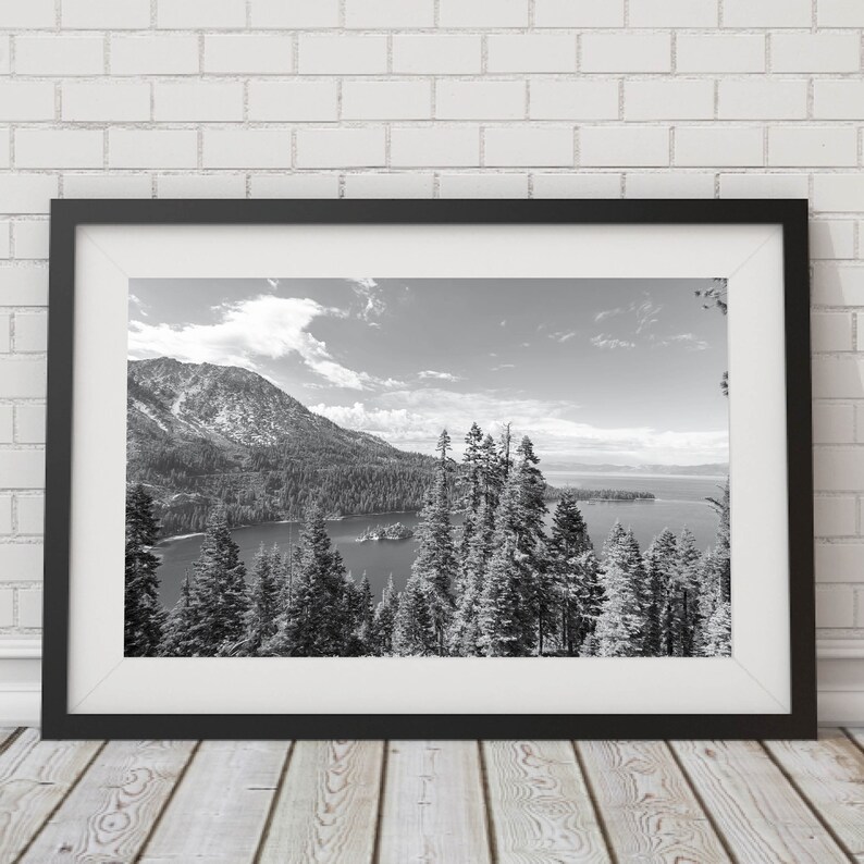 May include: A black and white photograph of a mountain lake with snow-capped mountains in the background. The lake is surrounded by trees and there is a small island in the middle of the lake.