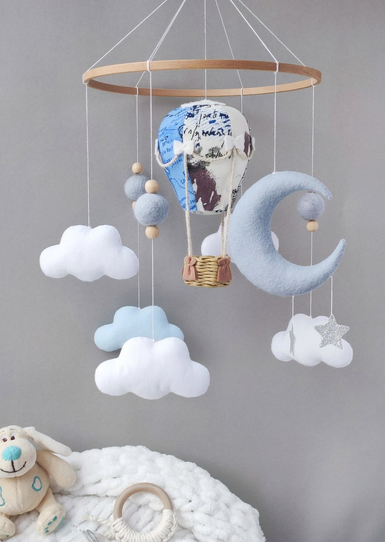 May include: A baby mobile with a blue and white hot air balloon, a blue crescent moon, white clouds, and a silver star. The mobile is made of felt and wood and is hanging from a wooden ring.