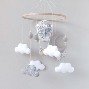 May include: A white and gray baby mobile with a hot air balloon, clouds, a crescent moon, and stars. The mobile is made of felt and wood.
