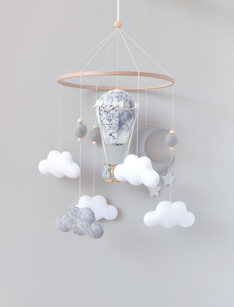 May include: A white and gray baby mobile with a hot air balloon, clouds, a crescent moon, and stars. The mobile is made of felt and wood.