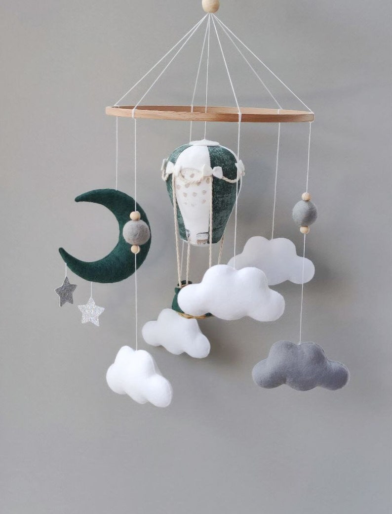 May include: A green and white hot air balloon, a crescent moon, and white and gray clouds hang from a wooden ring. The mobile is made of felt and is perfect for a baby's nursery.