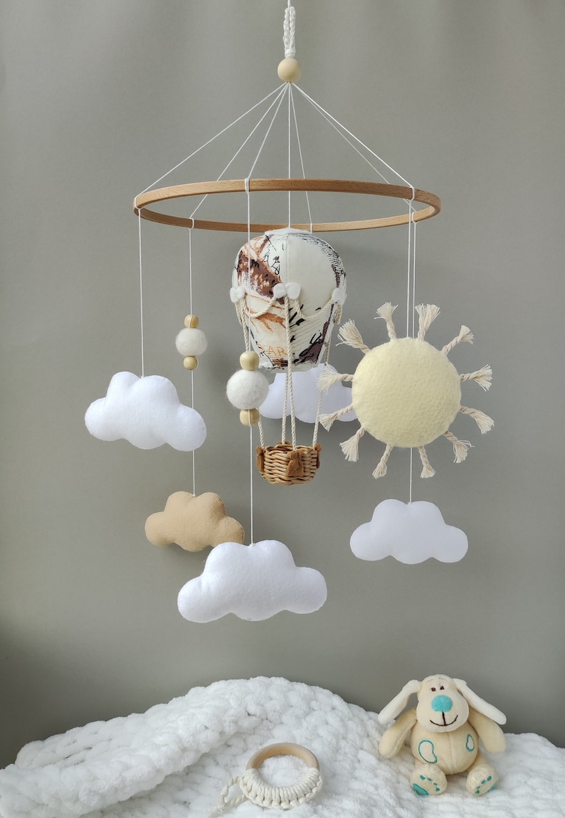 May include: A wooden baby mobile with a hot air balloon, clouds, a sun, and wooden beads. The mobile is hanging from a wooden ring.