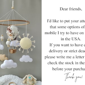 May include: A white, beige, and brown baby mobile with a hot air balloon, clouds, and a sun. The mobile is hanging from a wooden ring. The text "Dear friends, I'd like to put your attention that some options of this mobile I try to have on storage in the USA. If you want to have quick delivery or strict deadline please write me a letter and I'll check the stock in the USA before your purchase. Thank you!" is written on a white background.