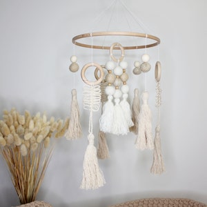 May include: A wooden baby mobile with white and beige felt balls, wooden rings, and macrame tassels. The mobile is hanging from a wooden hoop and is suspended by white string.