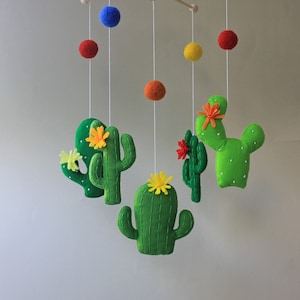 May include: A handmade baby mobile featuring felt cacti and colorful felt balls. The cacti are green with yellow and orange flower accents. The mobile is suspended from a wooden frame with a natural finish.