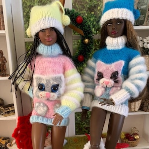Pu&ograve; includere: Due bambole che indossano maglioni di lana con disegni di gatti. Le bambole indossano cappelli di lana bianchi con pon pon e stivali di lana bianchi con pon pon. Le bambole sono sedute su una superficie di uncinetto verde e marrone.