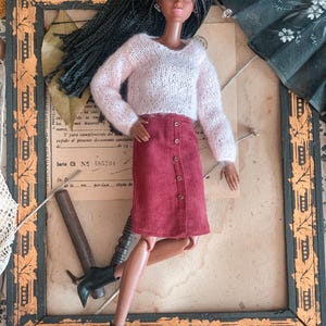 May include: A fashion doll in a light pink knitted sweater and a burgundy button-down skirt. The doll has long black hair and black ankle boots. The doll is posed in front of a vintage framed picture.