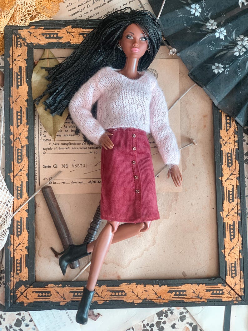 May include: A fashion doll in a light pink knitted sweater and a burgundy button-down skirt. The doll has long black hair and black ankle boots. The doll is posed in front of a vintage framed picture.