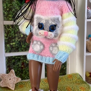 Pu&ograve; includere: Una bambola che indossa un maglione a righe rosa, blu e giallo con un design di gatto grigio. La bambola indossa anche un cappello di lana giallo e bianco e calzini di lana bianchi con pon pon.