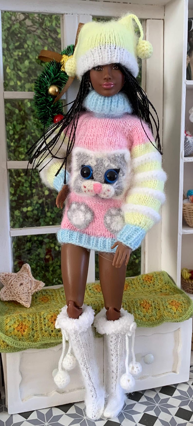 Pu&ograve; includere: Una bambola che indossa un maglione a righe rosa, blu e giallo con un design di gatto grigio. La bambola indossa anche un cappello di lana giallo e bianco e calzini di lana bianchi con pon pon.
