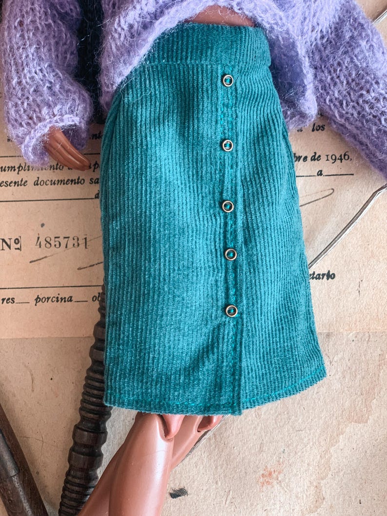 May include: A teal corduroy skirt with a button-down front. The skirt is paired with a light purple, fuzzy sweater. The background includes vintage paper with text and the number 485731.