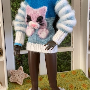 Pu&ograve; includere: Una bambola che indossa un maglione di lana blu, bianco e rosa con un design a gatto. La bambola indossa un cappello di lana bianco con un pon pon blu e stivali di lana bianchi con pon pon.