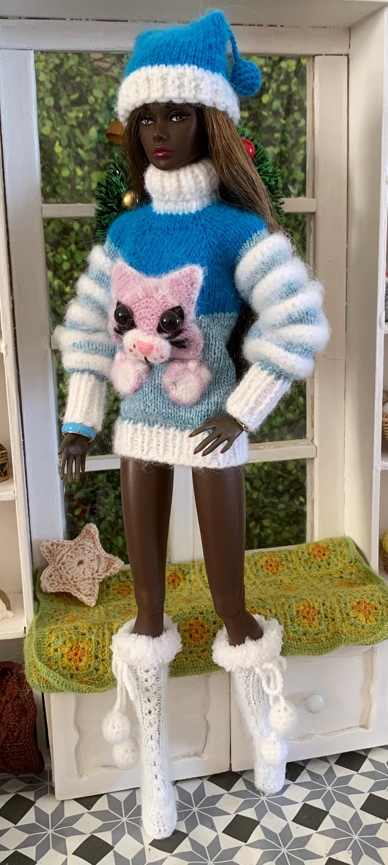 Pu&ograve; includere: Una bambola che indossa un maglione di lana blu, bianco e rosa con un design a gatto. La bambola indossa un cappello di lana bianco con un pon pon blu e stivali di lana bianchi con pon pon.