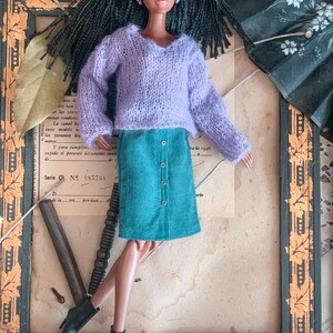 May include: A doll with dark hair and brown skin wearing a lavender sweater and a teal corduroy skirt with buttons. The doll is wearing black ankle boots. The doll is posed in front of a framed piece of paper.