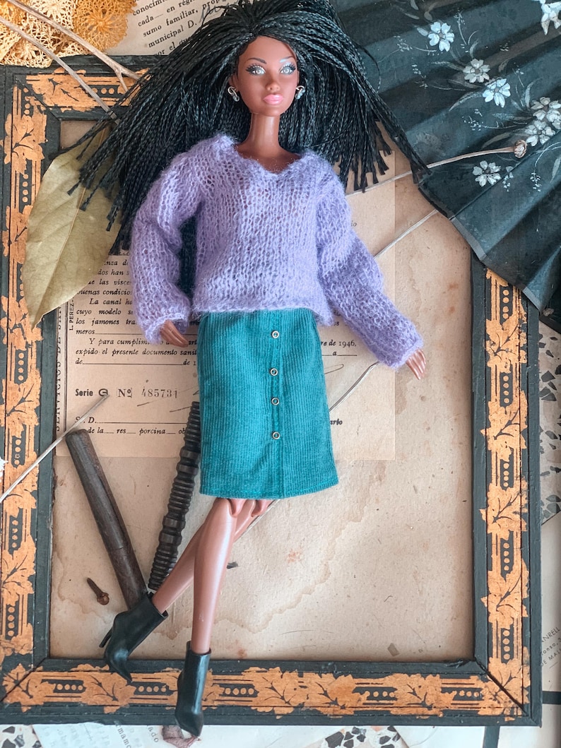 May include: A doll with dark hair and brown skin wearing a lavender sweater and a teal corduroy skirt with buttons. The doll is wearing black ankle boots. The doll is posed in front of a framed piece of paper.