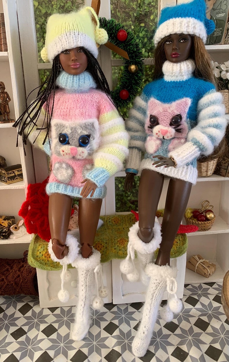Pu&ograve; includere: Due bambole che indossano maglioni di lana con disegni di gatti. Le bambole indossano cappelli di lana bianchi con pon pon e stivali di lana bianchi con pon pon. Le bambole sono sedute su una superficie di uncinetto verde e marrone.
