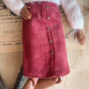 May include: A doll dressed in a white, fluffy jumper and a burgundy corduroy skirt. The skirt features a button-down design. The doll has brown skin and is wearing black boots.