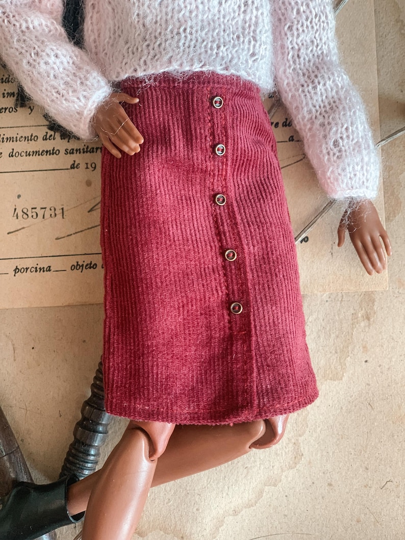 May include: A doll dressed in a white, fluffy jumper and a burgundy corduroy skirt. The skirt features a button-down design. The doll has brown skin and is wearing black boots.
