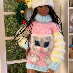 Pu&ograve; includere: Una bambola che indossa un maglione a righe rosa, giallo e blu con un viso di gattino grigio e impronte di zampe. La bambola indossa un cappello di lana giallo e bianco con un pon pon in cima.
