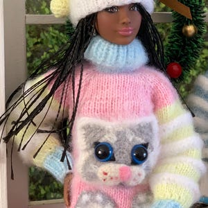 Pu&ograve; includere: Una bambola che indossa un maglione a righe rosa, giallo e blu con un viso di gatto grigio e impronte di zampe. La bambola indossa anche un cappello di lana giallo e bianco con un pon pon in cima.