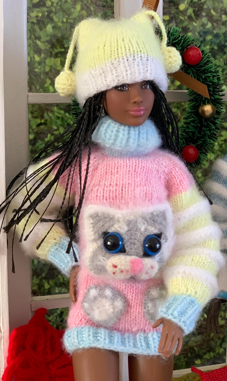 Pu&ograve; includere: Una bambola che indossa un maglione a righe rosa, giallo e blu con un viso di gatto grigio e impronte di zampe. La bambola indossa anche un cappello di lana giallo e bianco con un pon pon in cima.