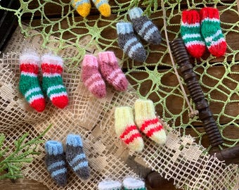 Può includere: Una collezione di calzini in miniatura lavorati a maglia in varie combinazioni di colori, tra cui strisce rosse e verdi, giallo e blu, rosa, grigio e crema con strisce rosse. Questi minuscoli calzini sono disposti a coppie su una rete.
