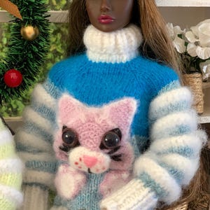 Pu&ograve; includere: Una bambola che indossa un maglione a righe blu e bianche con un'applicazione di gatto rosa. Il maglione ha un collo alto bianco e la bambola indossa un cappello di lana bianco.