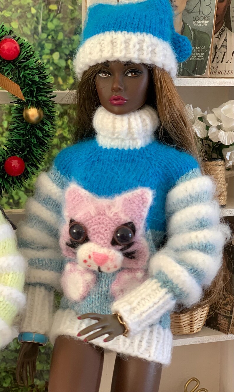 Pu&ograve; includere: Una bambola che indossa un maglione a righe blu e bianche con un'applicazione di gatto rosa. Il maglione ha un collo alto bianco e la bambola indossa un cappello di lana bianco.