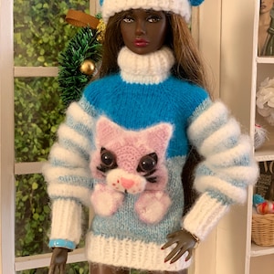 Pu&ograve; includere: Una bambola che indossa un maglione di lana blu e bianco con un design di gatto rosa. La bambola indossa anche un cappello di lana blu e bianco.
